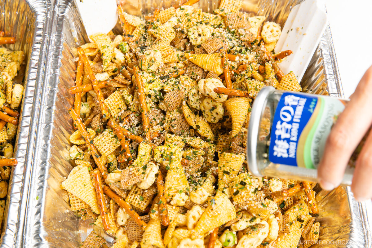 A hand sprinkles seasoning from a can over a tray of homemade snack mix containing pretzels, Chex cereal, crackers, and rice crackers, all laid out in a foil tray.