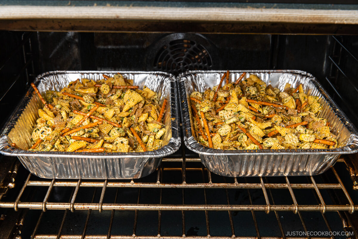 Two foil trays filled with seasoned Chex mix snacks bake in an oven. The mix includes cereal pieces, pretzel sticks, and seasoning, all distributed evenly in the trays.