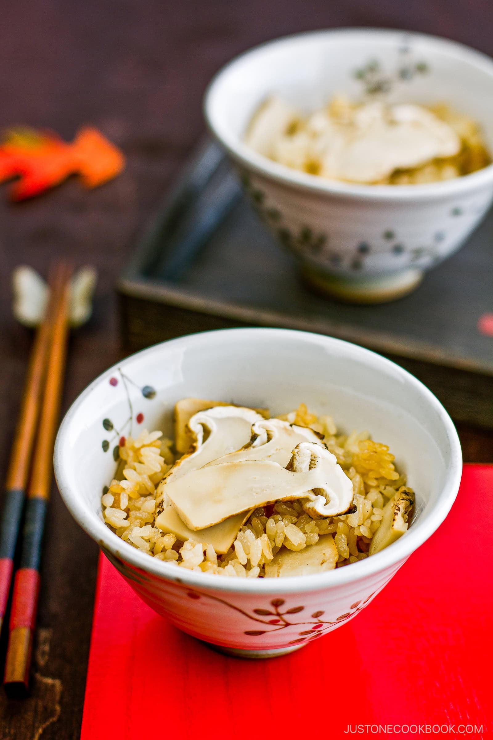 A bowl of rice topped with sliced mushrooms sits on a red mat, with another bowl and a pair of chopsticks in the background. The setting has a warm, rustic atmosphere.