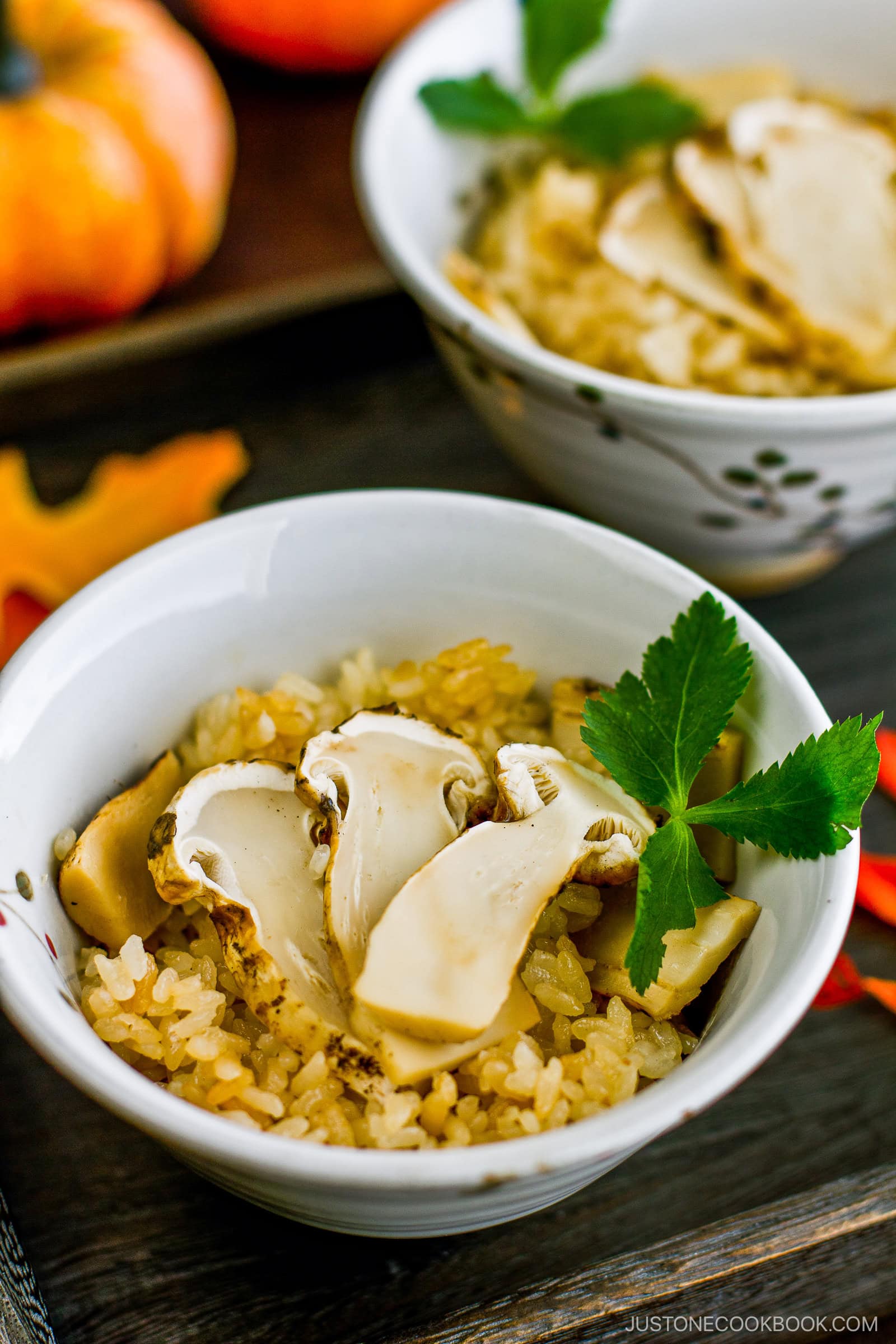 A bowl of rice topped with slices of mushroom and garnished with a sprig of green parsley, with another similar bowl and autumn decorations visible in the background.