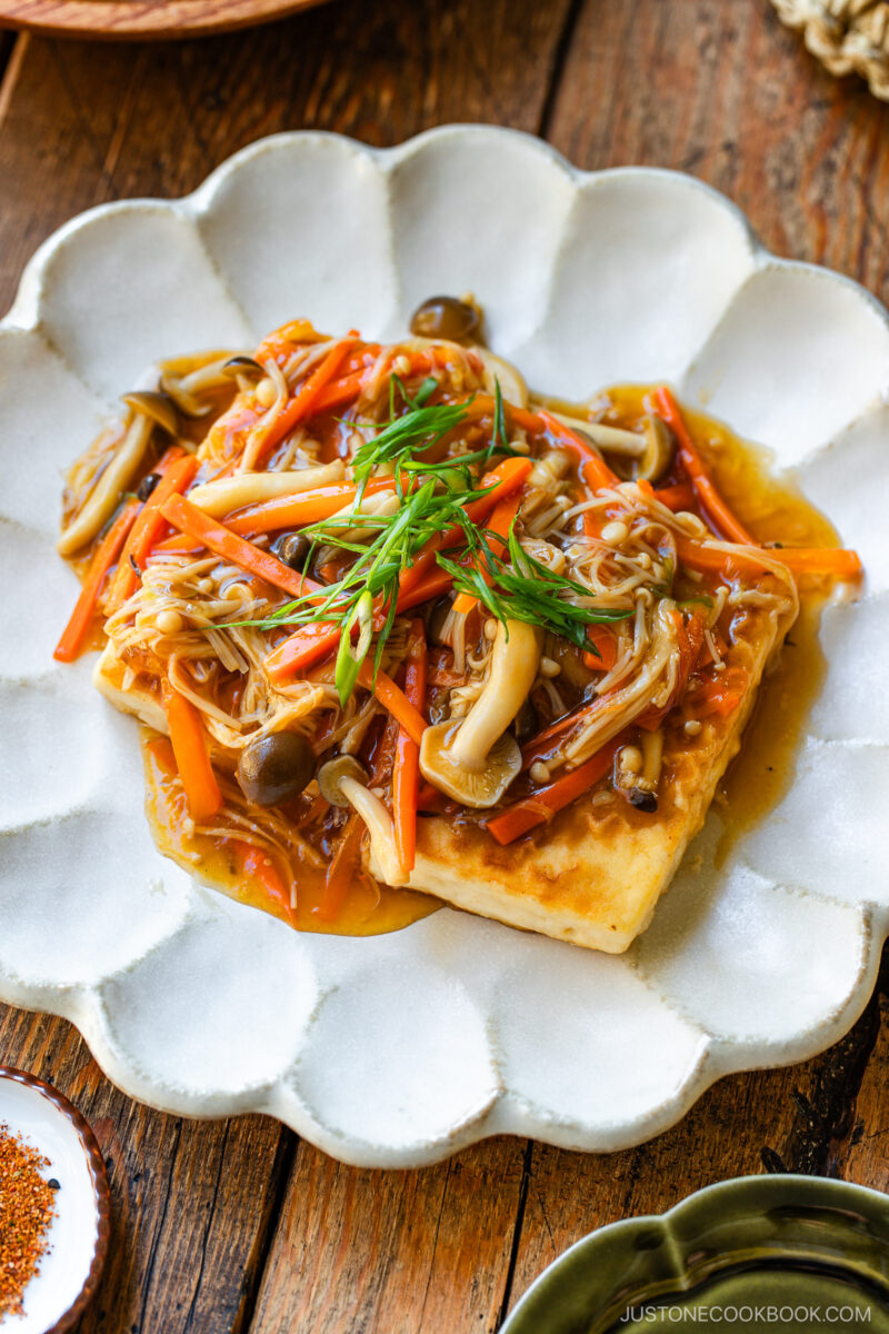 A plate of crispy tofu topped with stir-fried mushrooms, julienned carrots, and green onion, served with a savory brown sauce on a scalloped white dish.