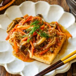 A square piece of tofu topped with stir-fried mushrooms, julienned carrots, and sauce, garnished with sliced green onions, served on a white scalloped plate next to chopsticks.