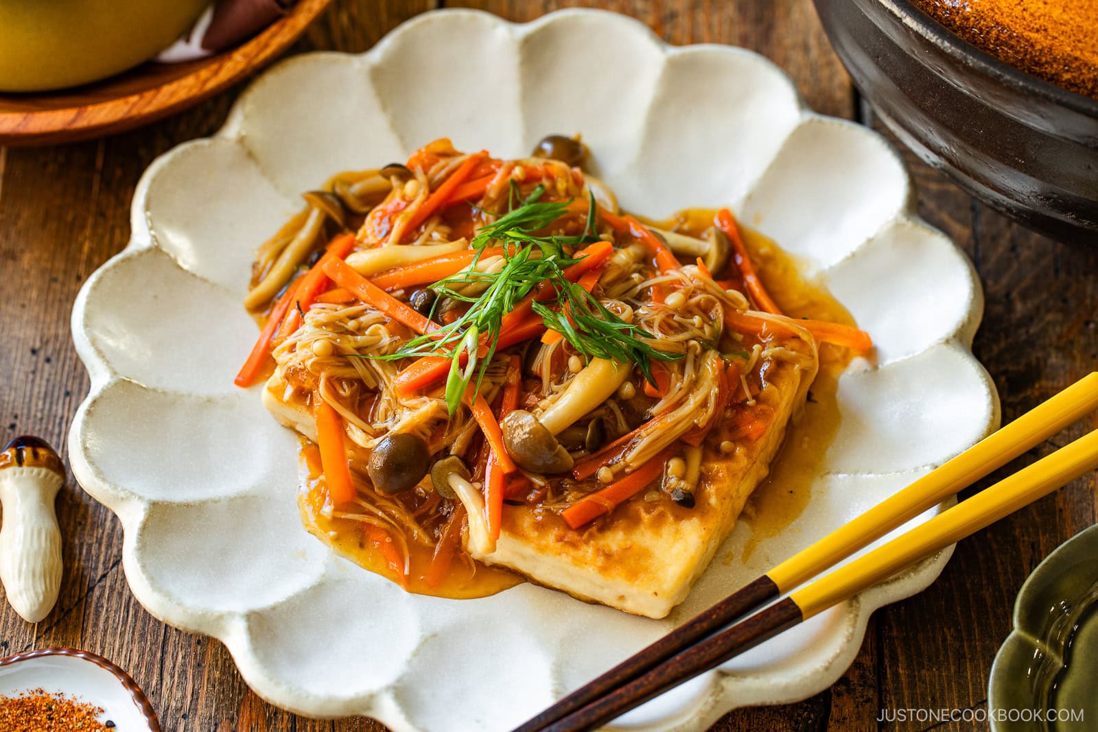 A square piece of tofu topped with stir-fried mushrooms, julienned carrots, and sauce, garnished with sliced green onions, served on a white scalloped plate next to chopsticks.