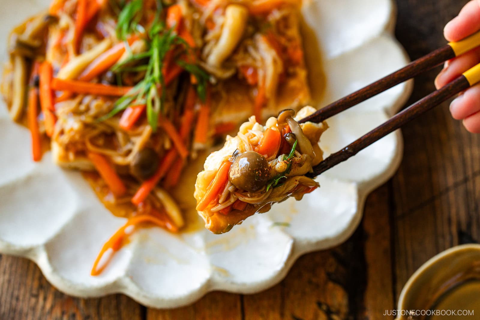 Chopsticks holding a bite of tofu topped with colorful sautéed vegetables and mushrooms, with more tofu and vegetables on a white plate in the background.