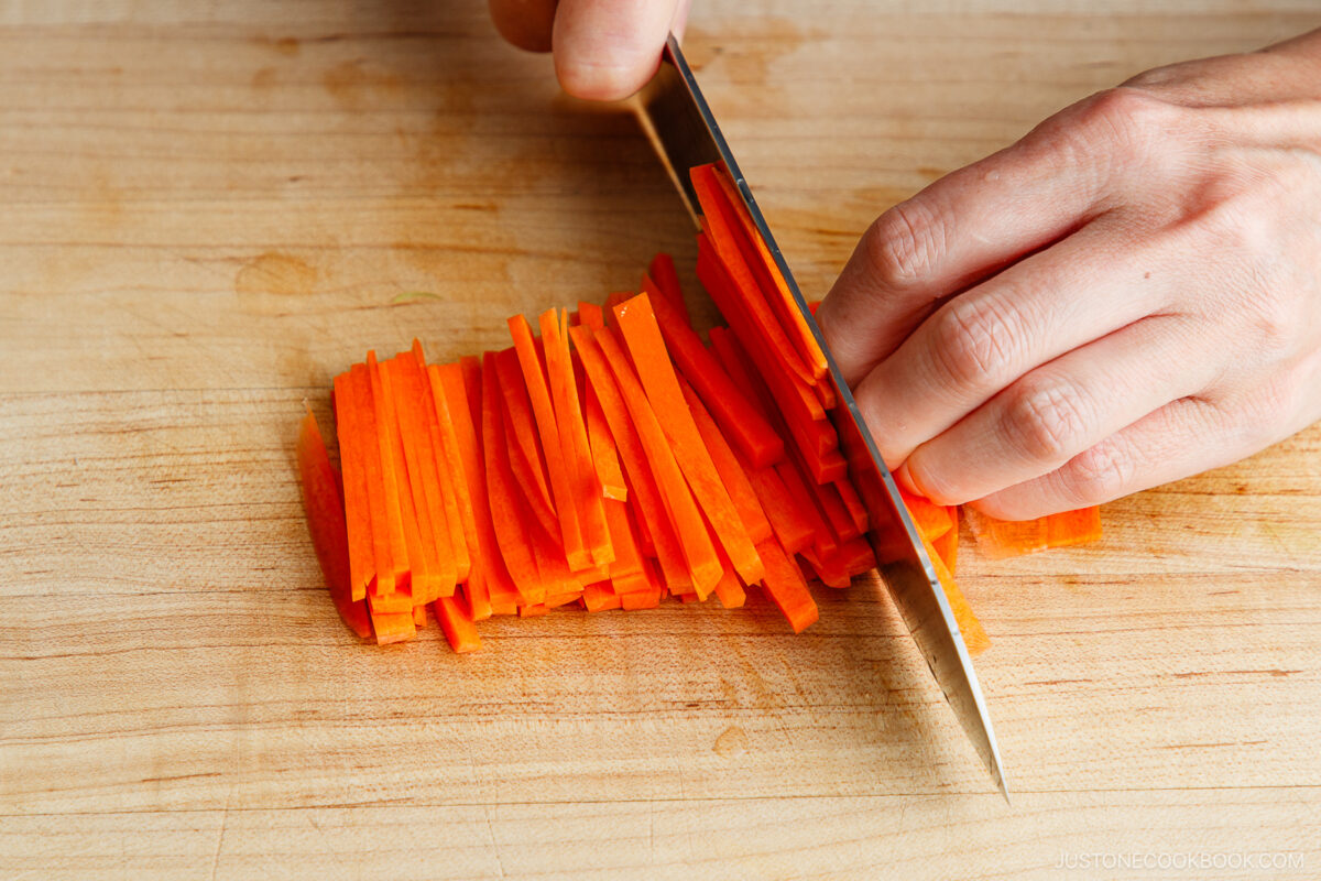 A hand uses a knife to cut carrots into thin matchstick-like strips on a wooden cutting board.