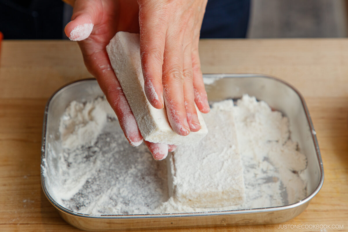 A person dusting rectangular pieces of fish with flour in a metal tray, preparing them for cooking. Their hands are covered in flour, and a wooden surface is visible underneath the tray.