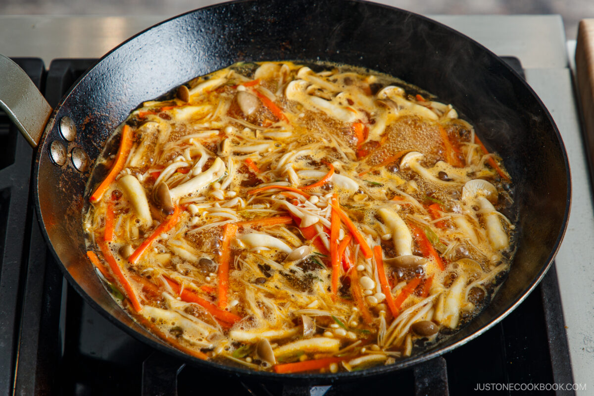 A pan of hot and sour soup simmers on a stovetop, filled with sliced mushrooms, shredded carrots, and other vegetables in a flavorful broth.
