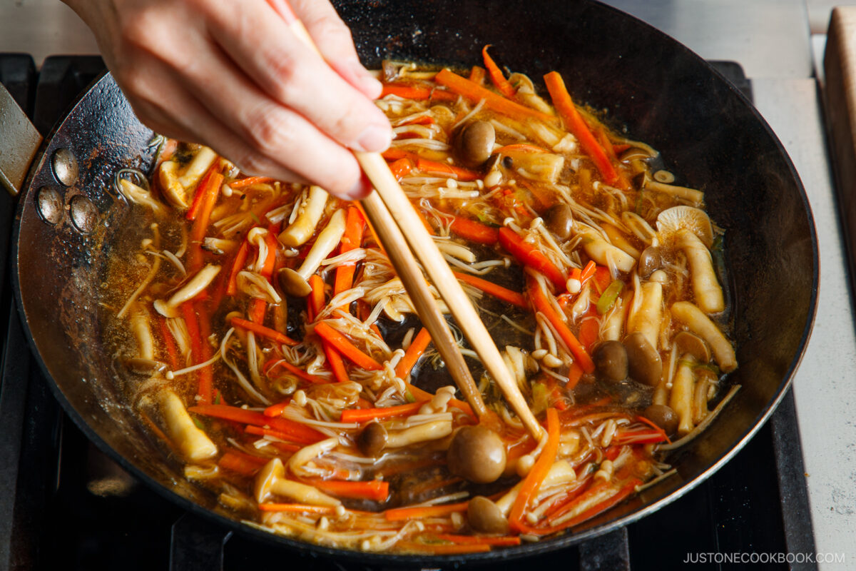 A hand uses chopsticks to stir a pan filled with mushrooms and julienned carrots simmering in a brown broth on a stovetop.