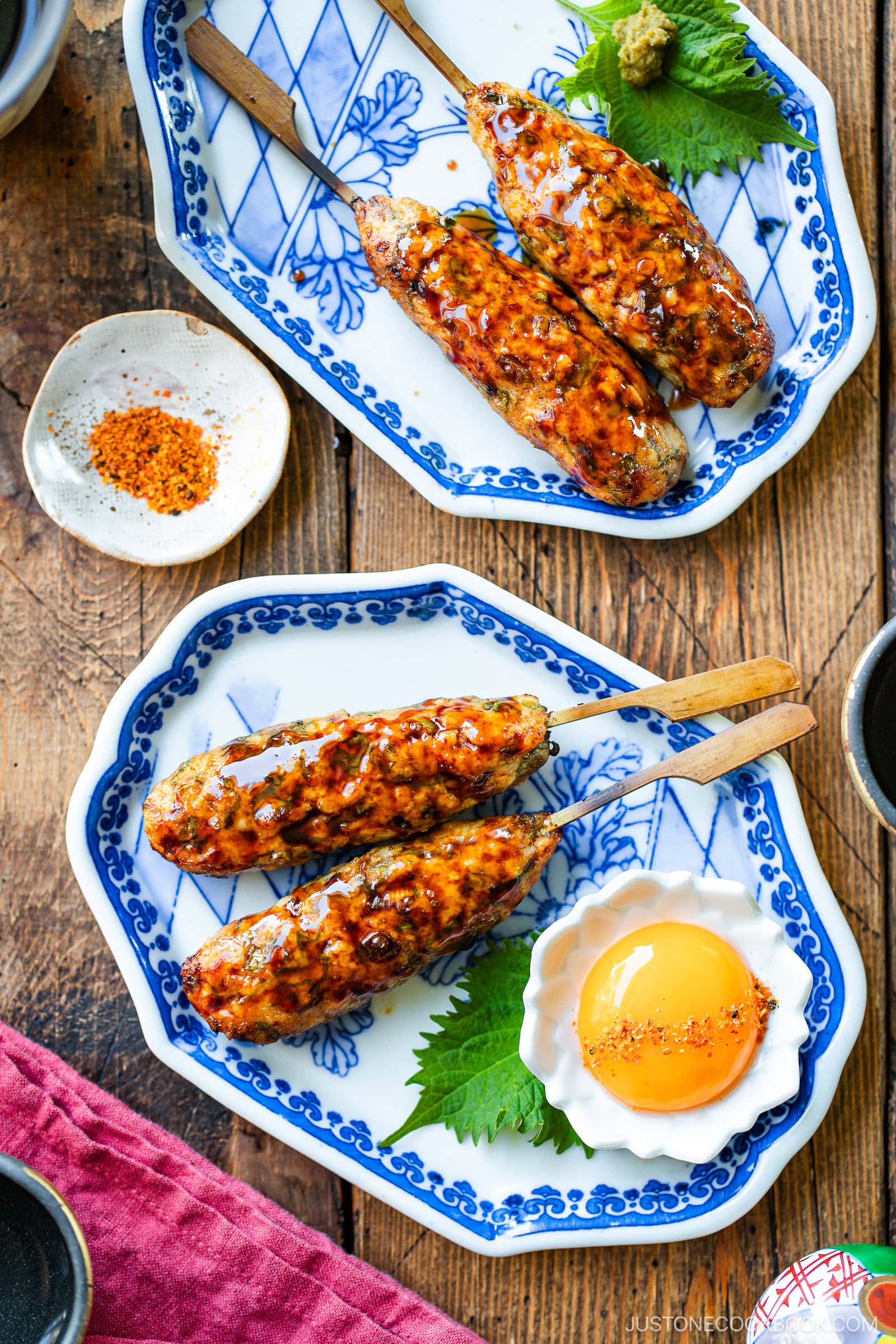 Two blue-and-white plates each hold skewered grilled meat glazed with sauce. One plate has three skewers with a raw egg yolk and shiso leaf, the other has two skewers. Condiments and a red napkin are nearby on a wooden table.