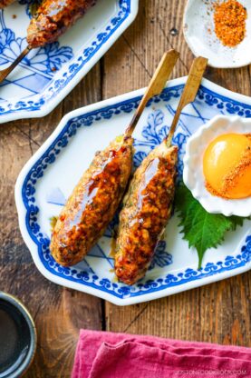 Two skewers of grilled chicken tsukune glazed with sauce on a patterned plate, served with a raw egg yolk, a green shiso leaf, and a small dish of spice on a wooden table.