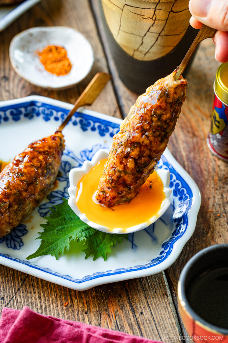 Grilled minced meat skewers glazed with sauce, one being dipped into a small dish of raw egg yolk, served on a blue and white plate with a green shiso leaf on a rustic wooden table.