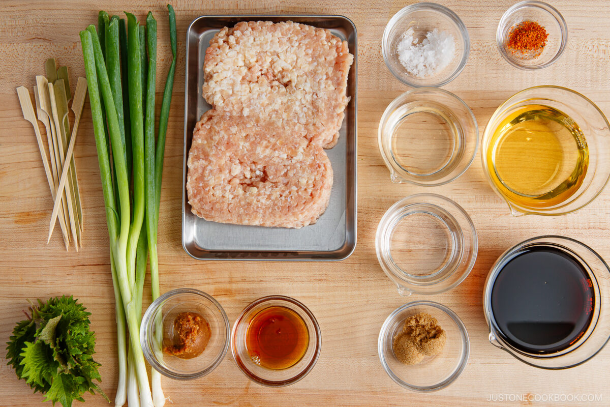 Top-down view of chopped green onions, skewers, shiso leaves, ground chicken, and small bowls of various sauces, seasonings, and spices arranged neatly on a wooden surface.