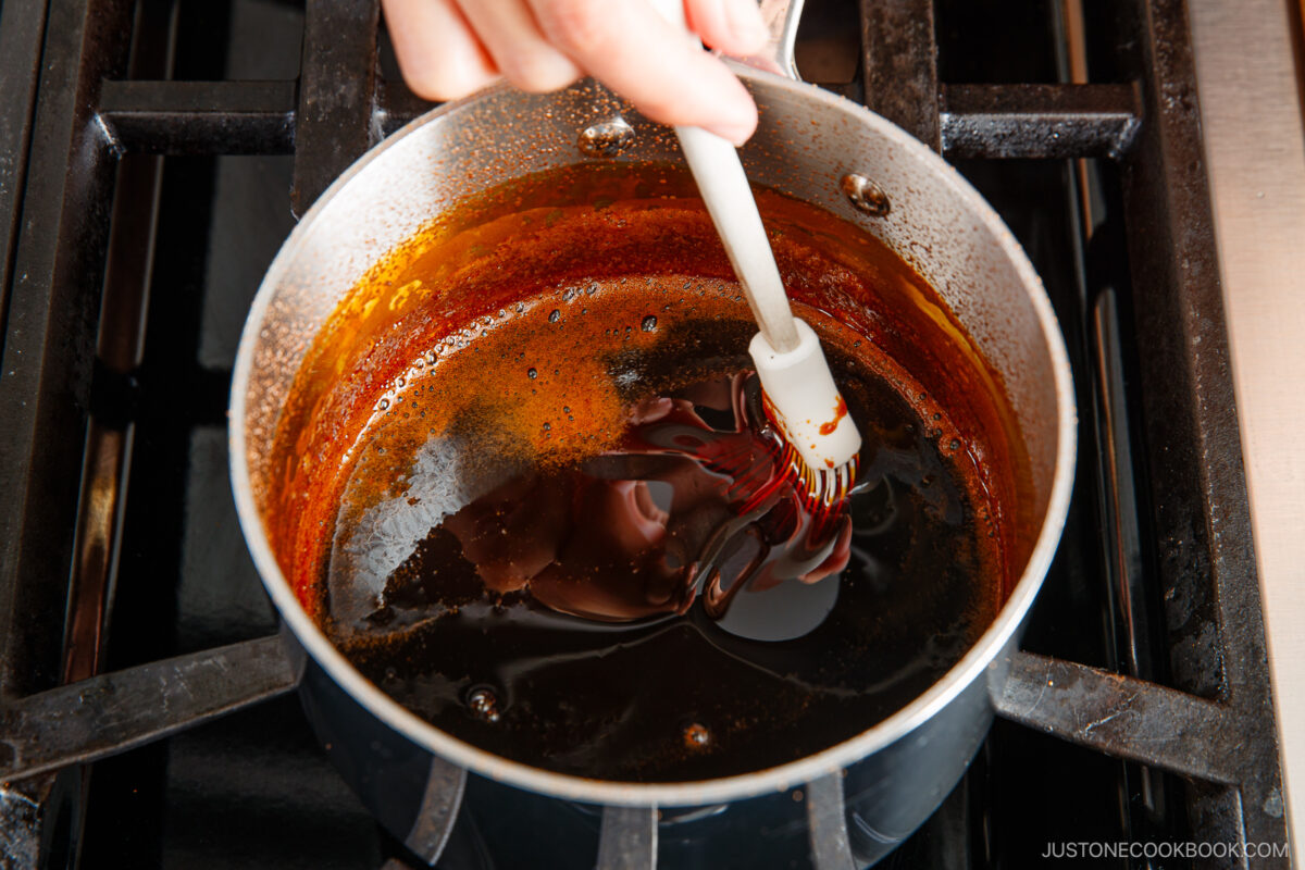 A hand stirs a dark, glossy sauce in a saucepan with a white spatula on a stovetop.