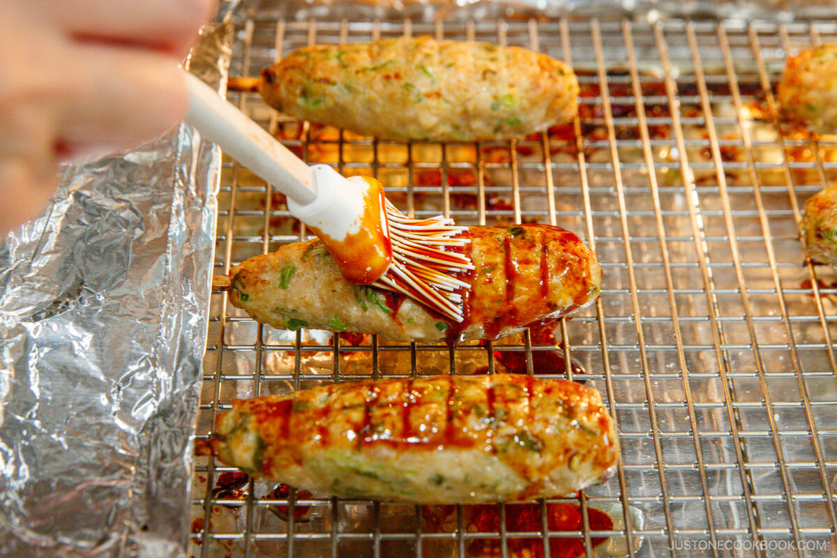A hand uses a brush to apply sauce to oval-shaped grilled meat patties on a wire rack lined with foil.