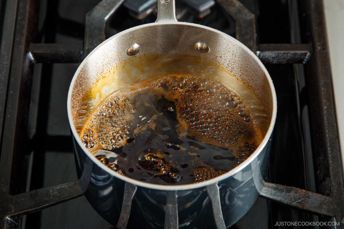 A metal saucepan with bubbling dark liquid, possibly a sauce or syrup, simmers on a gas stove burner.