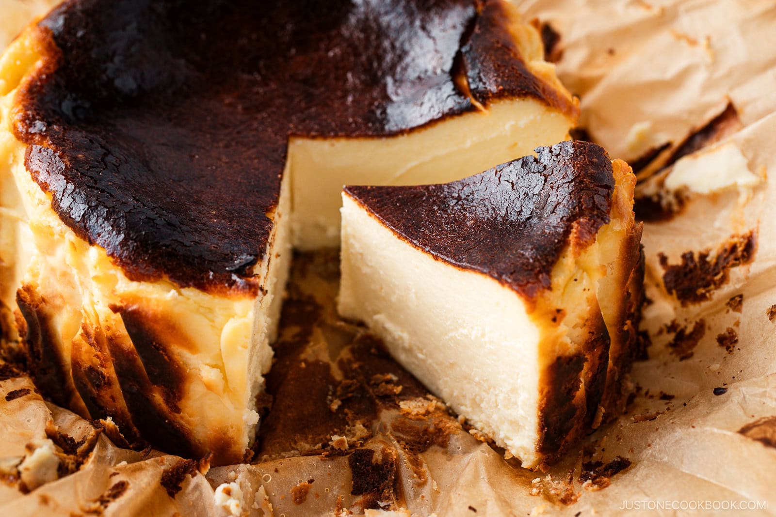 A close-up of a Basque burnt cheesecake on crinkled parchment paper, with a single slice cut and slightly pulled out, showing its creamy interior and caramelized top.