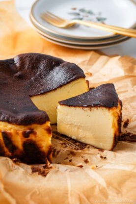 A sliced Basque burnt cheesecake sits on parchment paper, with its creamy interior and caramelized top visible. Plates and a fork are stacked in the background.