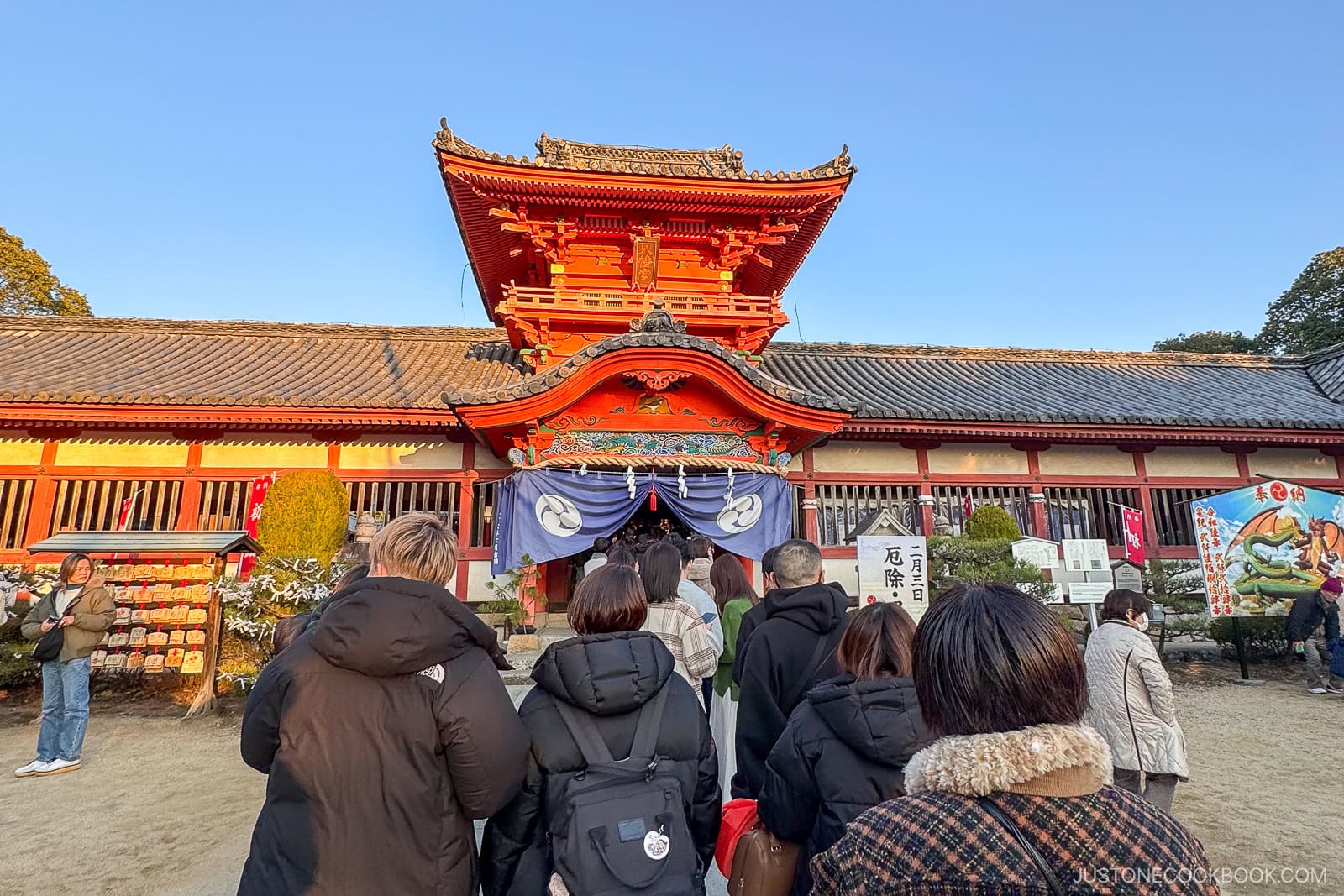 A group of people stand in line outside a traditional Japanese shrine with ornate red architecture, under a clear blue sky, likely during a festival or special event.