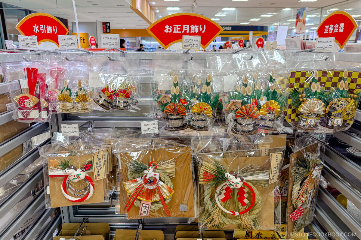 Shelves in a Japanese store display New Year’s decorations, including colorful ornaments made of paper, straw, pine, and bamboo, with red and gold accents and signs in Japanese above each section.