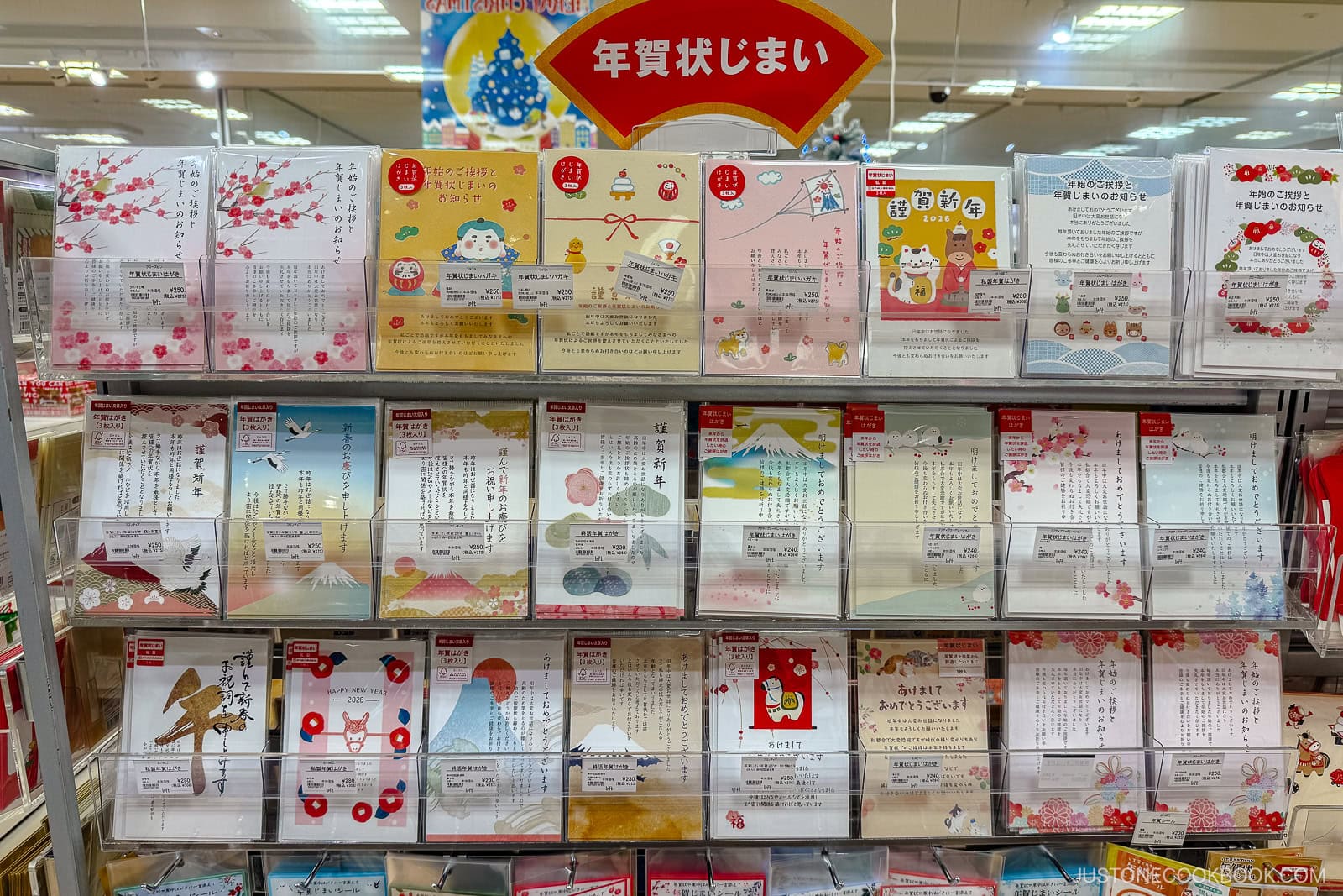 Display of colorful New Year greeting cards with Japanese designs and illustrations, organized on store shelves under a red sign with Japanese text. Cards feature flowers, animals, Mt. Fuji, and festive motifs.