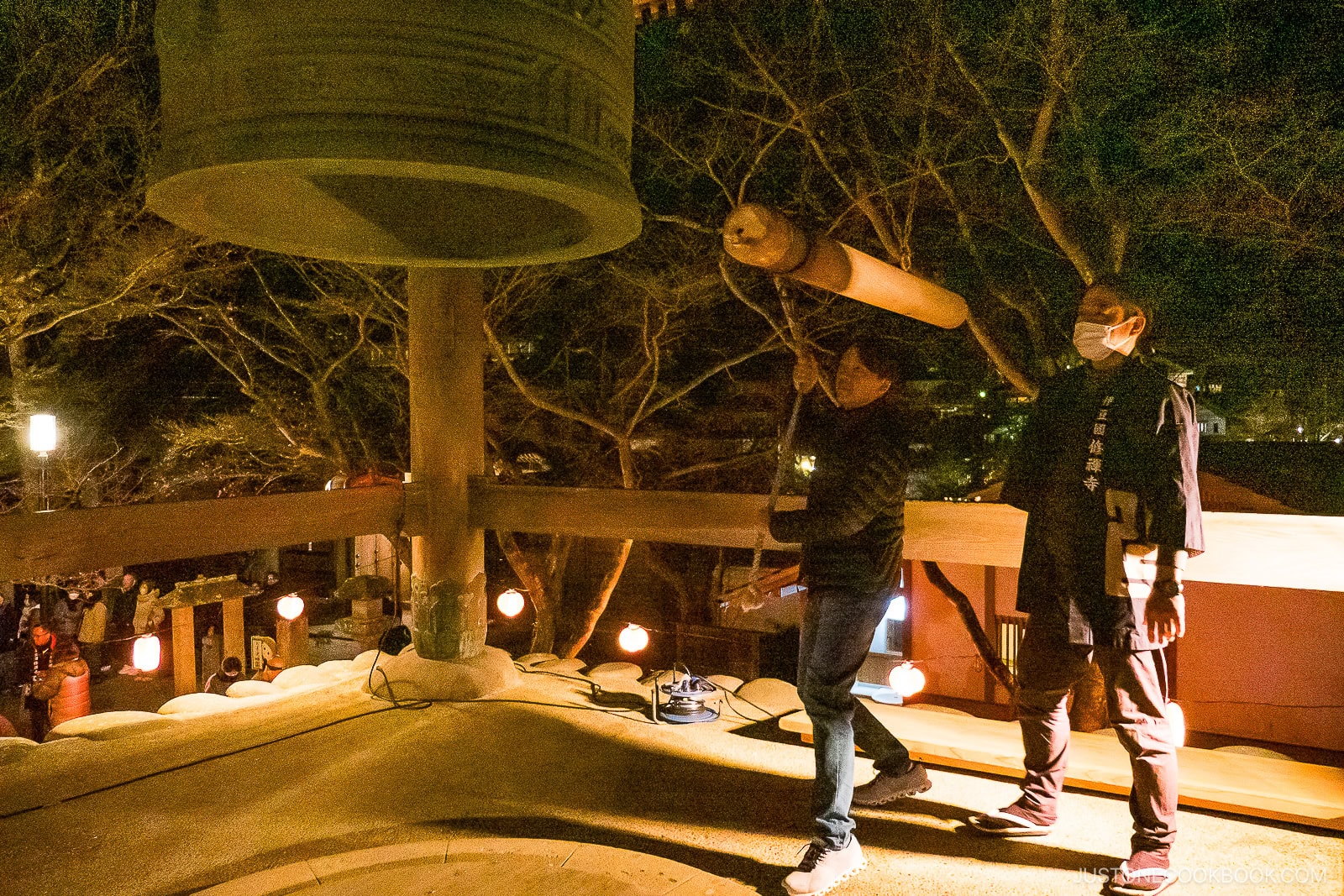 Two people at night, one swinging a wooden beam to strike a large bronze bell, while the other stands nearby. Lanterns and trees are visible in the background, creating a festive and traditional atmosphere.