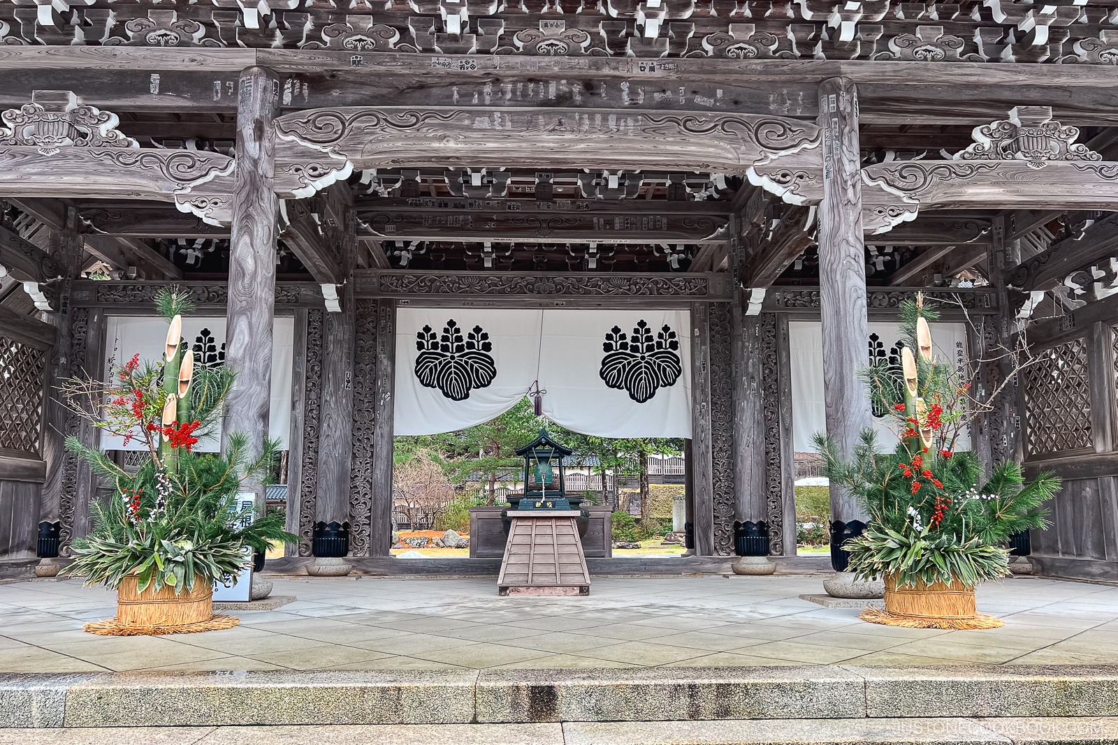 A traditional Japanese temple entrance decorated with large straw-bottomed arrangements of pine, bamboo, and red berries, known as kadomatsu, for the New Year. White banners with black crests hang in the background.