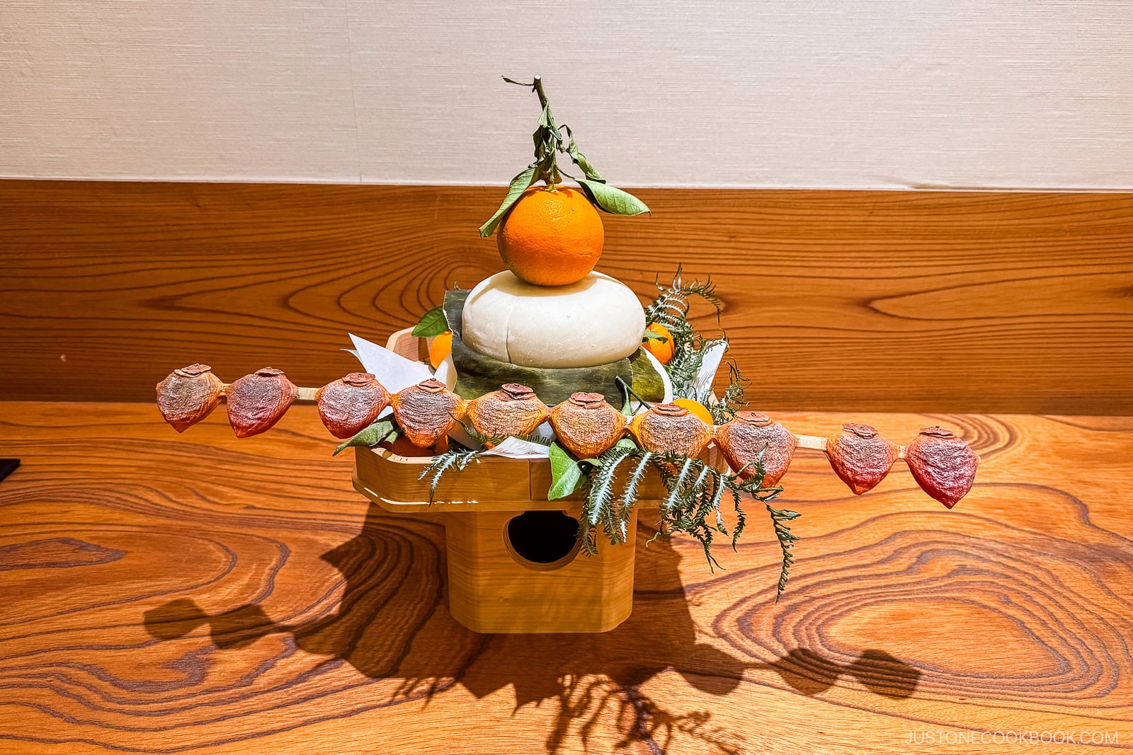 A traditional Japanese New Year kagami mochi display with two stacked rice cakes, a mandarin orange on top, and decorative elements, set on a wooden surface.