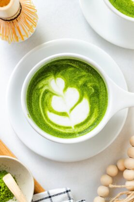 A cup of matcha latte with white leaf-shaped latte art on top, placed on a saucer. Nearby are a bamboo whisk, a bowl of matcha powder, and wooden beads, all arranged on a white surface.