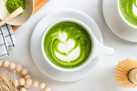 A top view of a white cup filled with matcha latte, featuring latte art on the frothy green surface. Surrounding items include a whisk, beads, a cloth napkin, and a bowl of matcha powder.