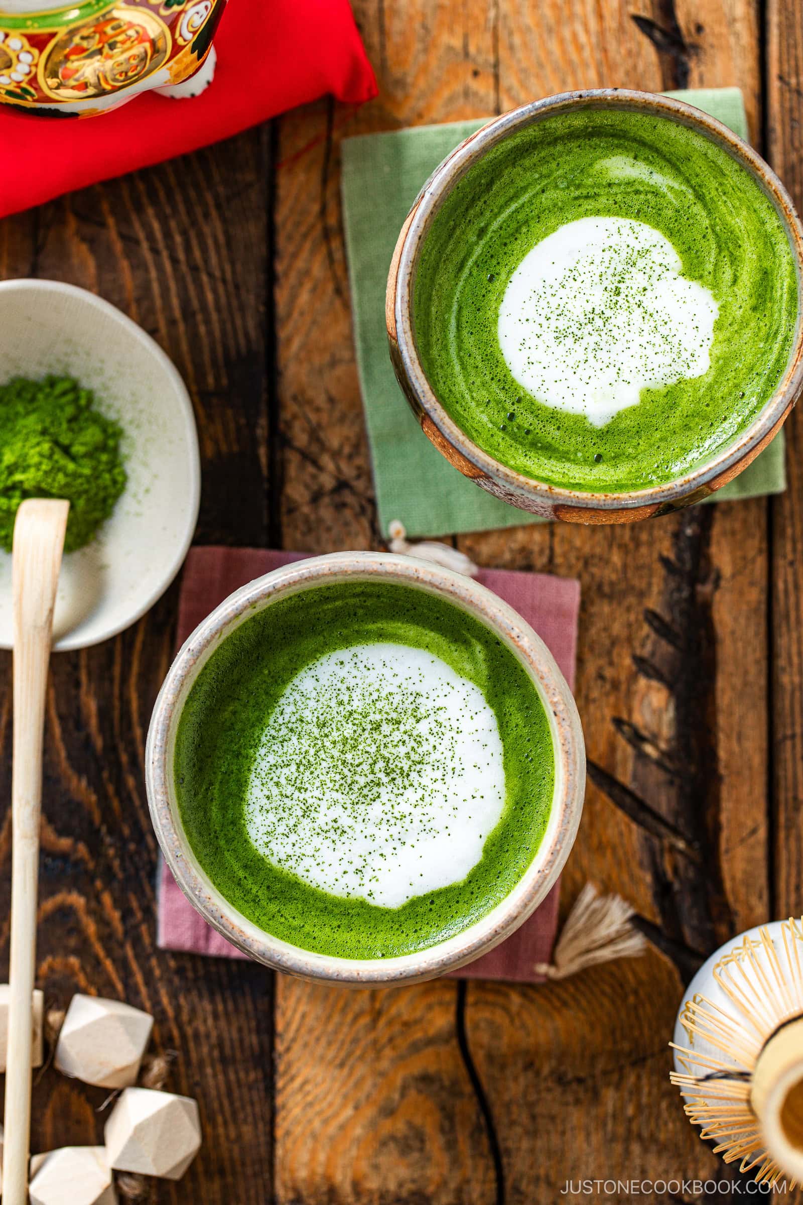 Two bowls of vibrant green matcha latte with white foam on top sit on a rustic wooden table, surrounded by a bowl of matcha powder, a bamboo whisk, a spoon, and decorative napkins.