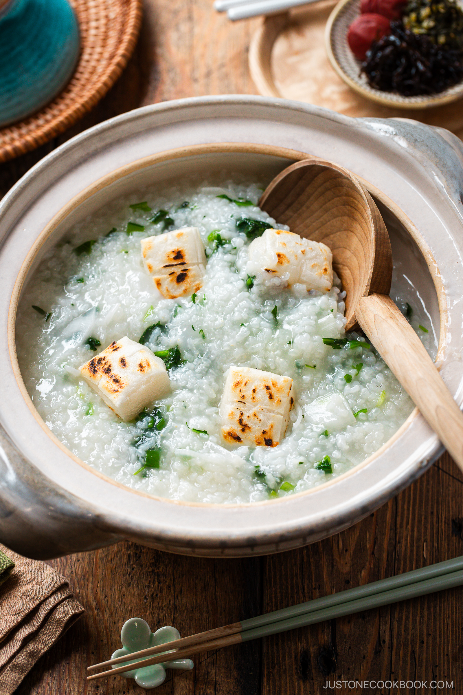 A bowl of Japanese rice porridge topped with grilled mochi cubes and chopped green onions, served in a clay pot with a wooden spoon on a wooden table. Chopsticks and small side dishes are nearby.