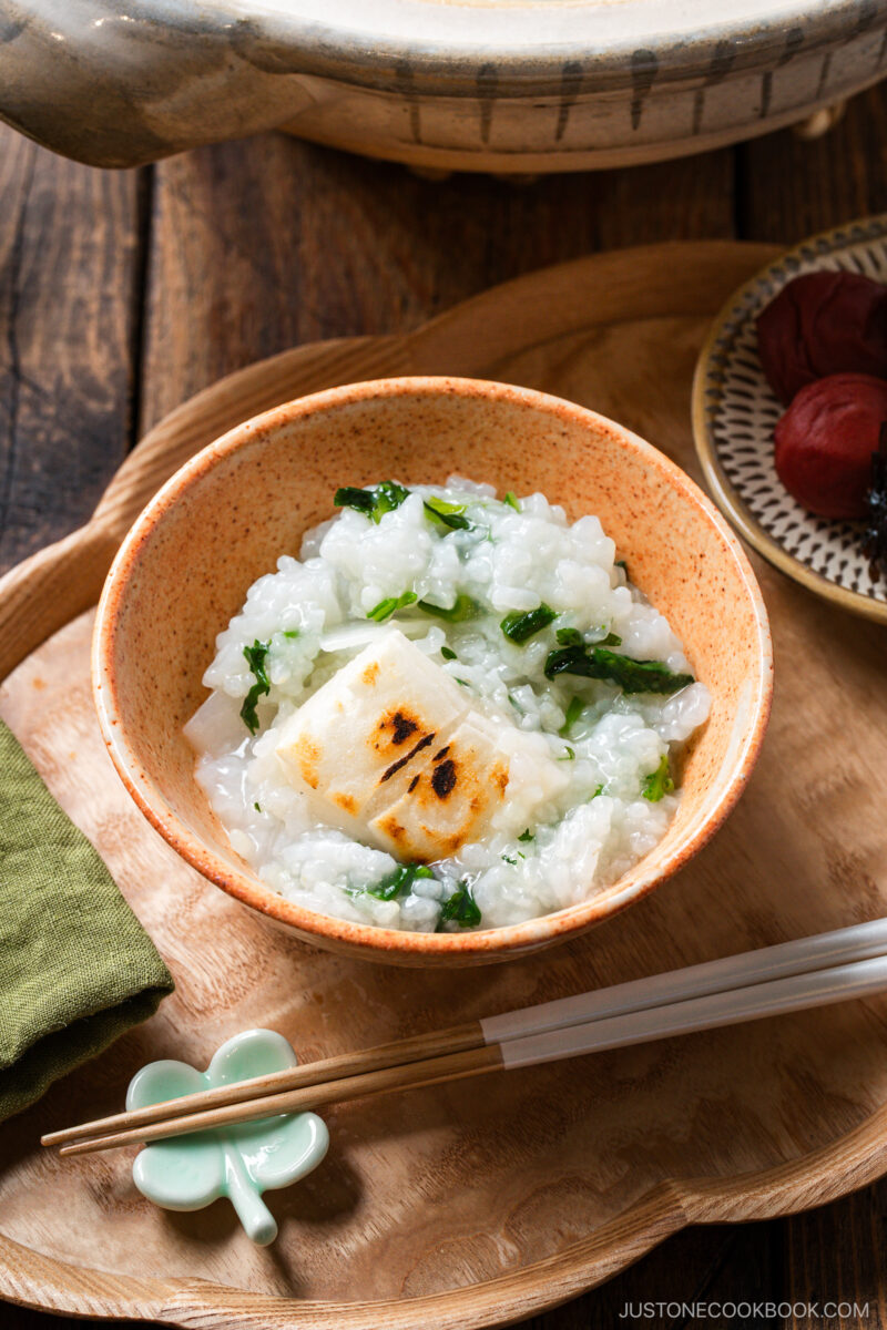A bowl of rice porridge with leafy greens and a piece of grilled mochi on top, served on a wooden tray with chopsticks, a chopstick rest, and a small plate of pickled plums on the side.