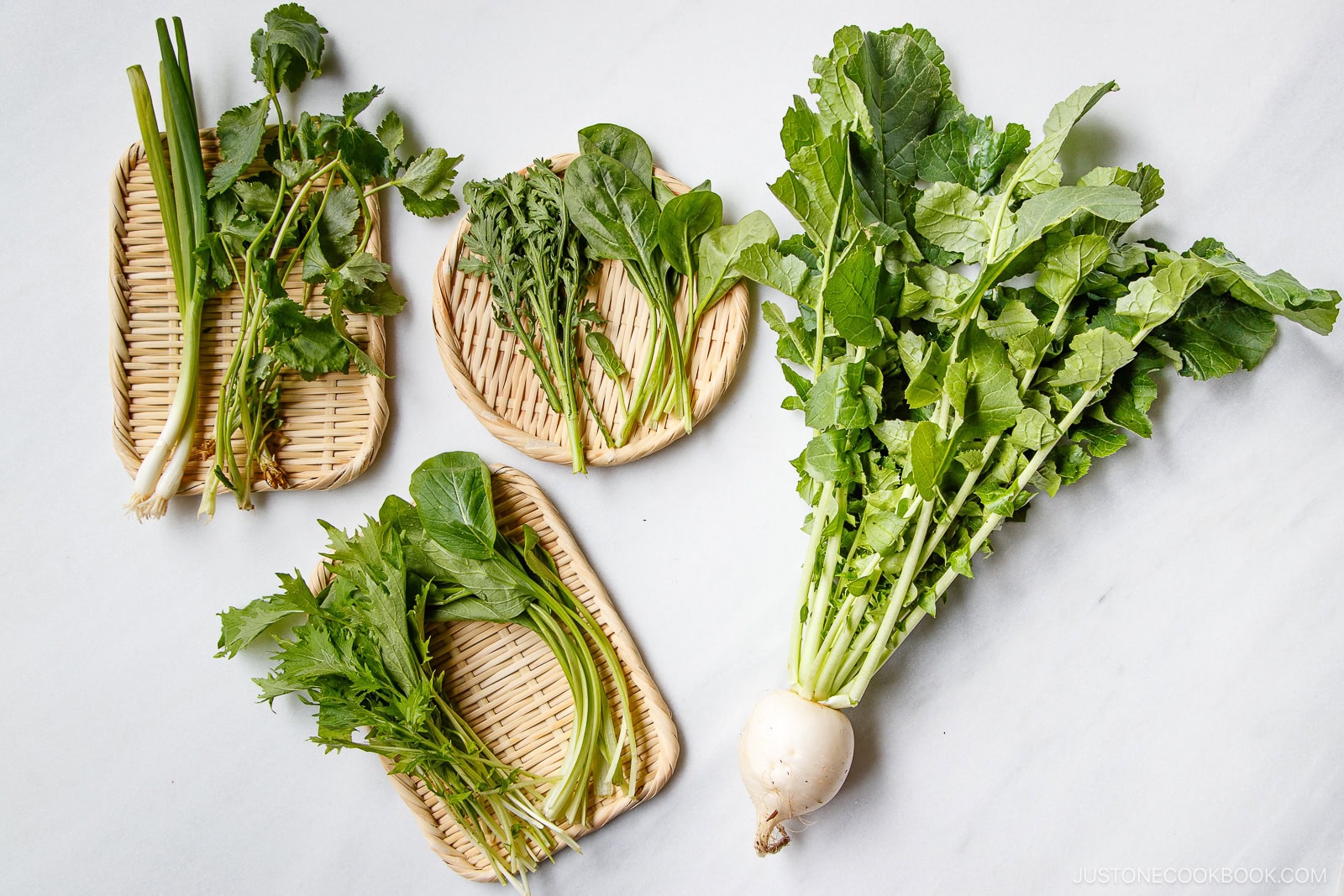 Five types of Japanese leafy greens, including turnip greens and others, are arranged on small woven bamboo trays and a marble surface. The greens are fresh with long stems and vibrant leaves.