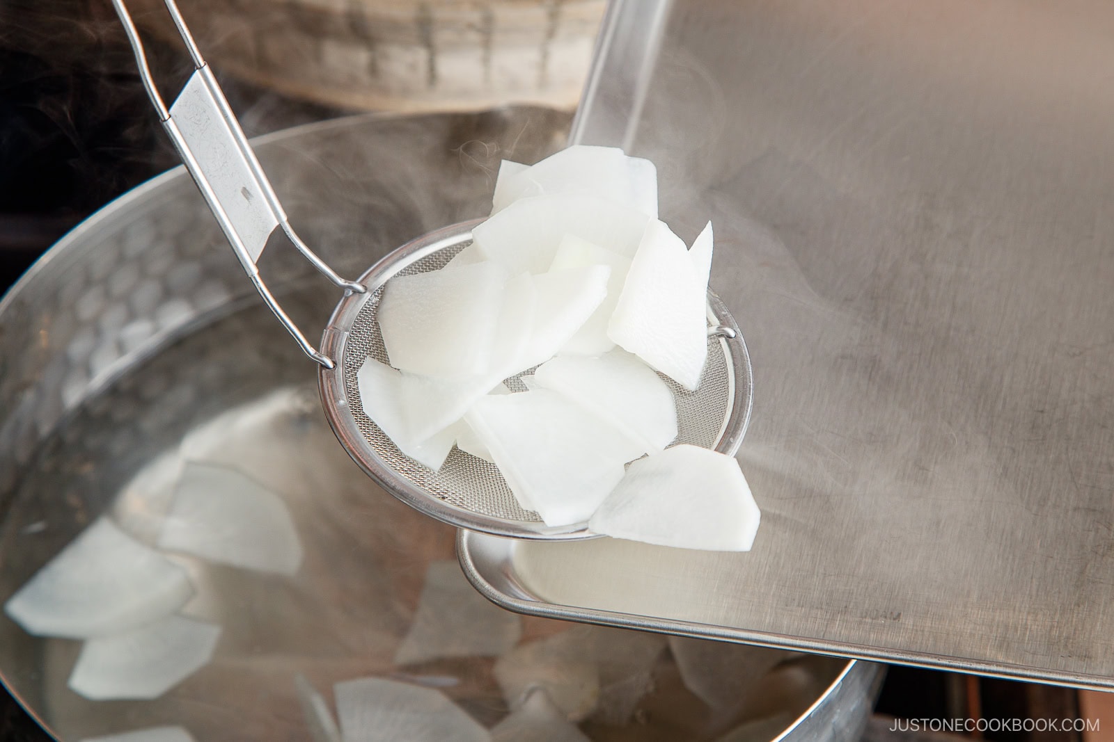 Thinly sliced white daikon pieces being lifted from a pot of hot water with a mesh strainer, next to a metal tray. Steam is rising from the pot.