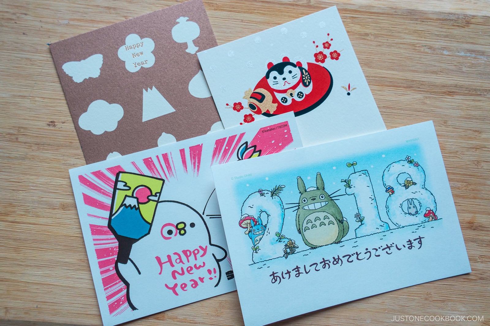 Four colorful Japanese New Year cards with cute illustrations, including a mountain, a cat, a character holding a fan, and Totoro with 2018 in the background, laid out on a wooden surface.