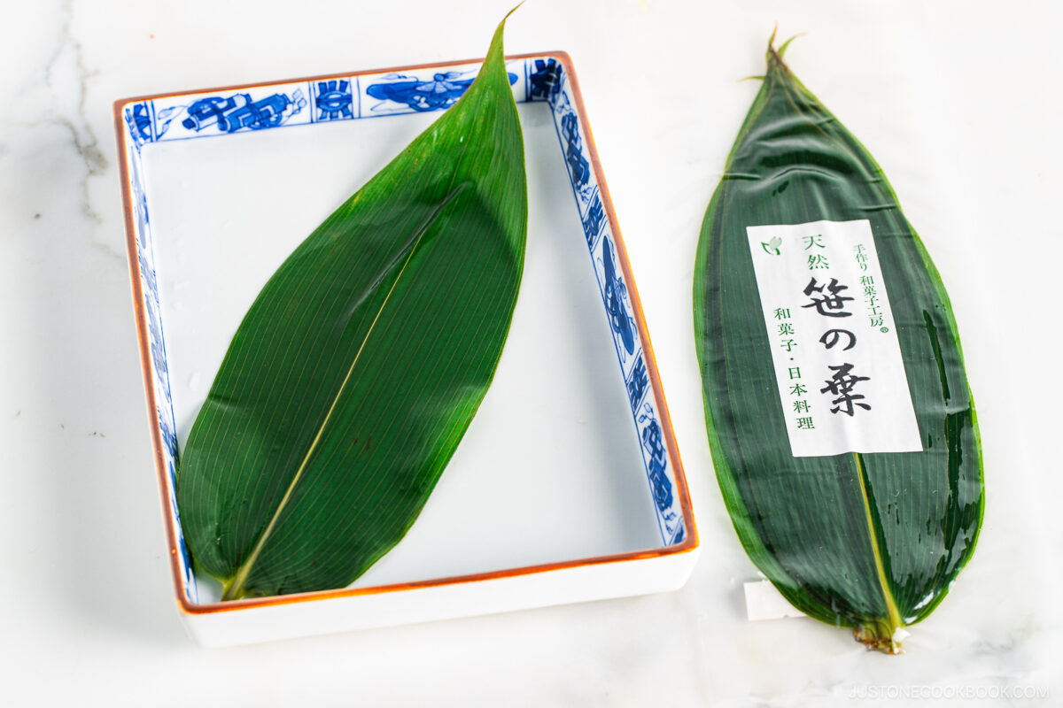 A rectangular dish filled with clear jelly topped with a large green bamboo leaf, next to another bamboo leaf wrapped in plastic packaging with Japanese text, on a white surface.