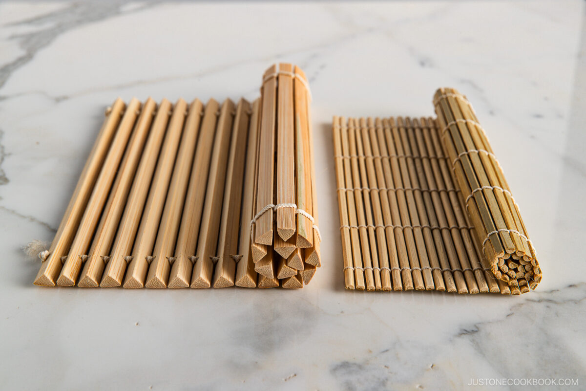 Two bamboo sushi rolling mats, one made of flat bamboo strips (left) and one of round bamboo sticks (right), are placed side by side on a white marble surface.