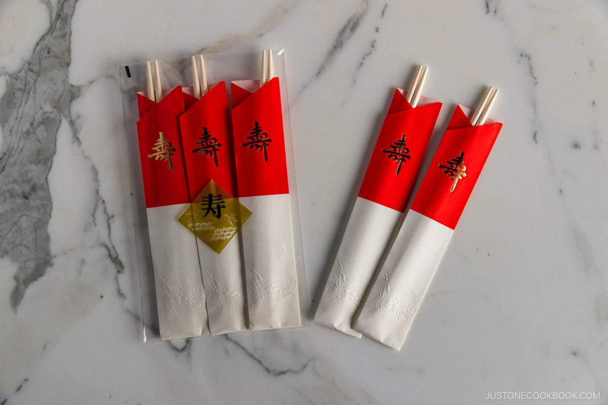 Four chopstick sets in white and red paper sleeves with gold and red kanji, plus two loose sets, placed on a light marble surface. The packaging has decorative patterns and a gold seal.