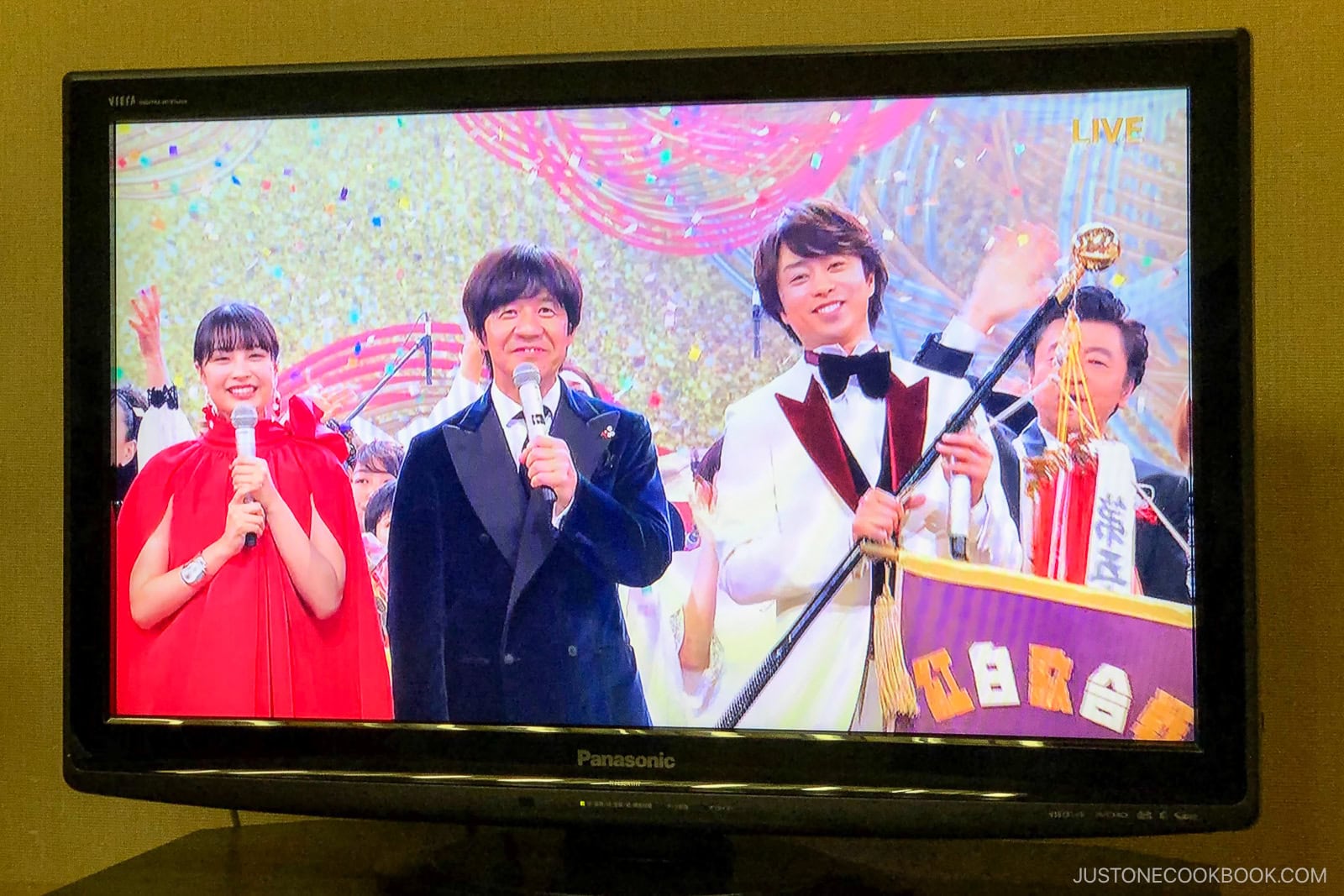 A television displays a Japanese awards show with three smiling hosts in formal attire—one woman in red, two men in suits, one holding a trophy—standing on a colorful, festive stage.