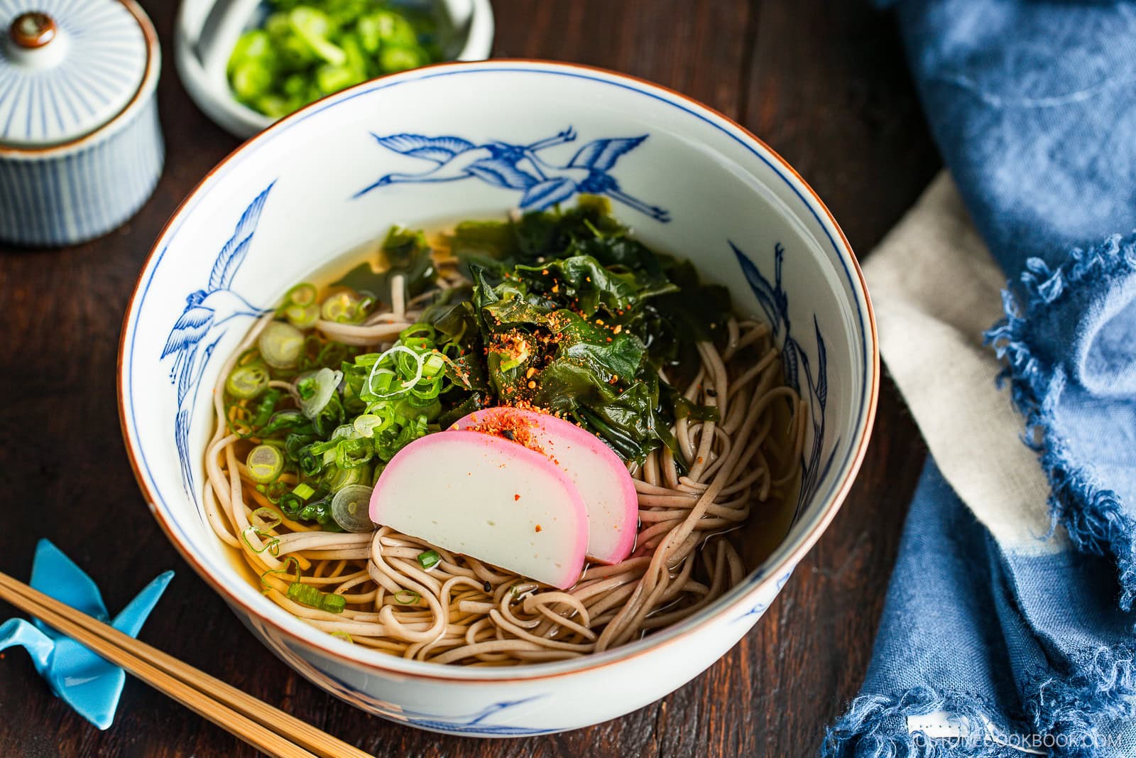 A bowl of soba noodle soup topped with sliced fish cake, seaweed, chopped green onions, and seasoning, with chopsticks and a blue napkin beside it on a wooden table.