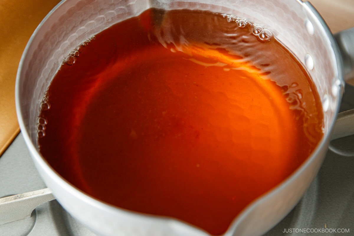 Close-up of a small metal saucepan containing a translucent amber-colored liquid, likely a type of broth or sauce, sitting on a stove.