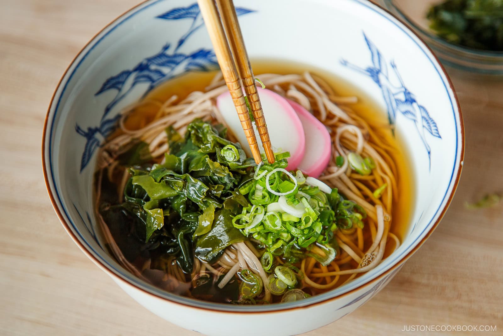 A bowl of Japanese soba noodle soup topped with sliced green onions, wakame seaweed, and fish cake, with chopsticks lifting noodles from the broth.