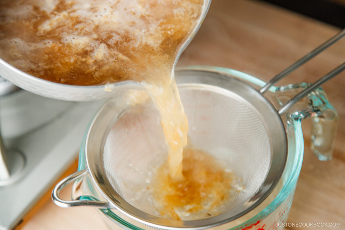 Hot broth is being poured from a pot through a fine mesh strainer into a glass measuring cup, separating solids from the liquid on a kitchen countertop.