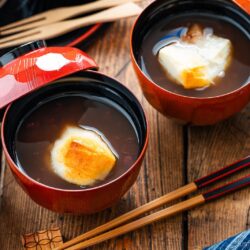 Two bowls of sweet red bean soup topped with grilled mochi sit on a wooden table, accompanied by a cup of green tea, chopsticks, and a blue-striped cloth.