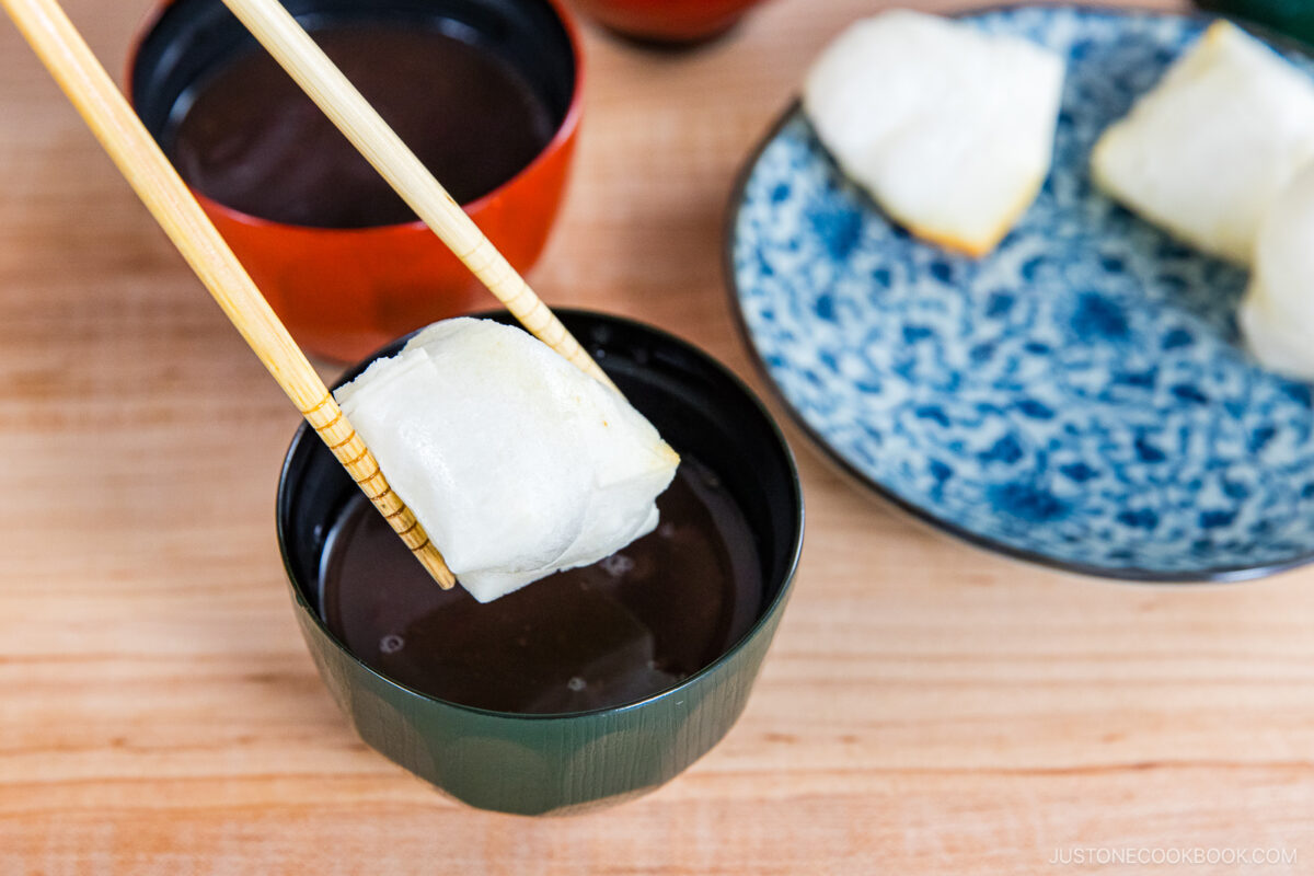 A pair of chopsticks holds a piece of toasted mochi over a bowl of sweet red bean soup, with more mochi and soup bowls visible on a wooden table.