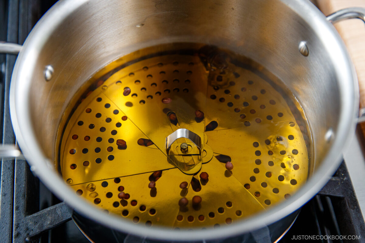 A pot of hot oil sits on a stove with a metal popcorn popper insert and several popcorn kernels at the bottom, ready to pop.