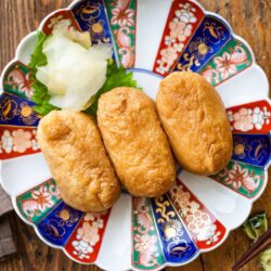 Three pieces of inari sushi on a decorative Japanese plate with colorful patterns, garnished with pickled ginger and a green leaf, placed on a wooden table with chopsticks and a small cup nearby.
