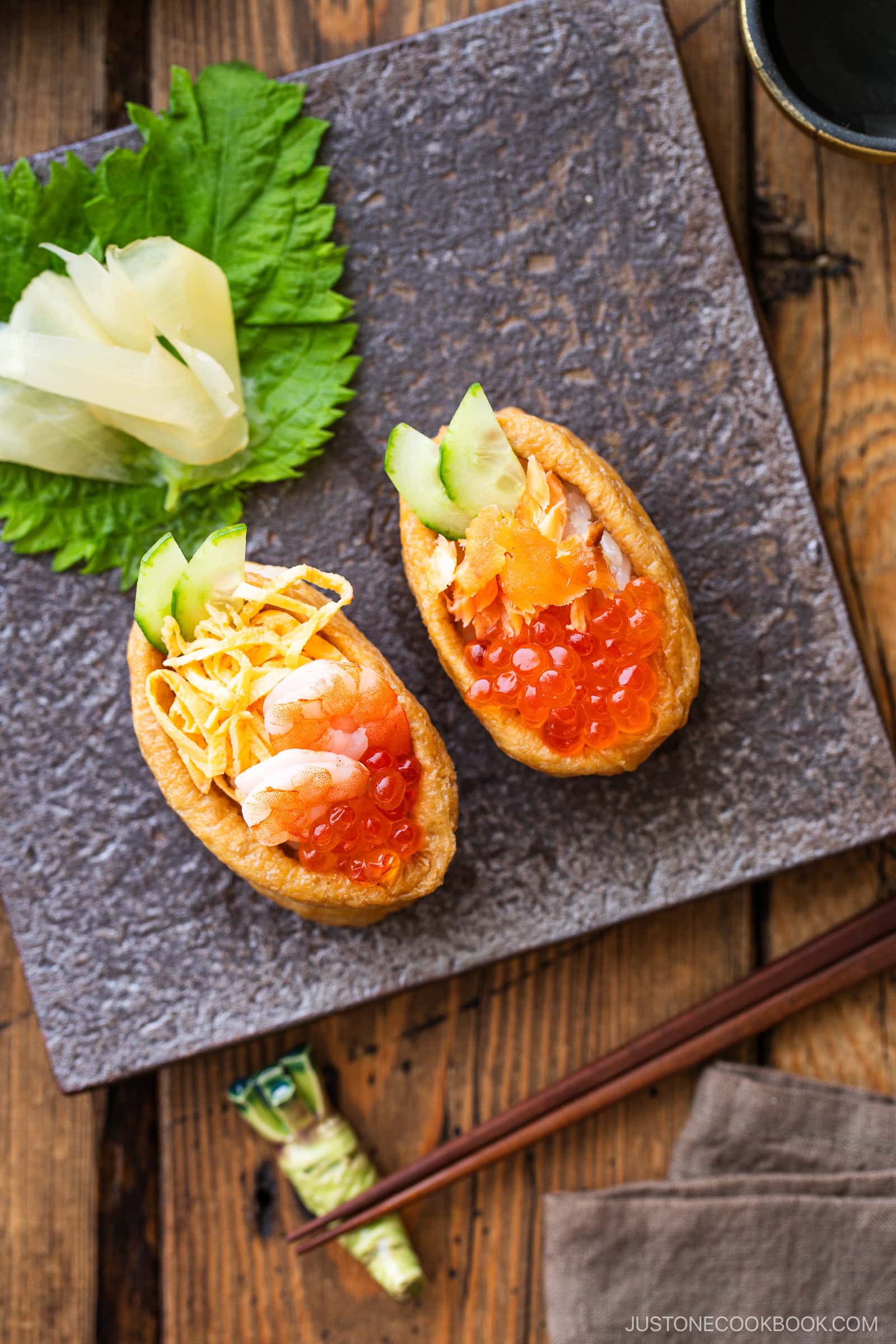 Two inari sushi pockets filled with rice, shrimp, cucumber, egg, and salmon roe on a square stone plate. Garnished with pickled ginger and a green shiso leaf, with chopsticks and wasabi nearby.