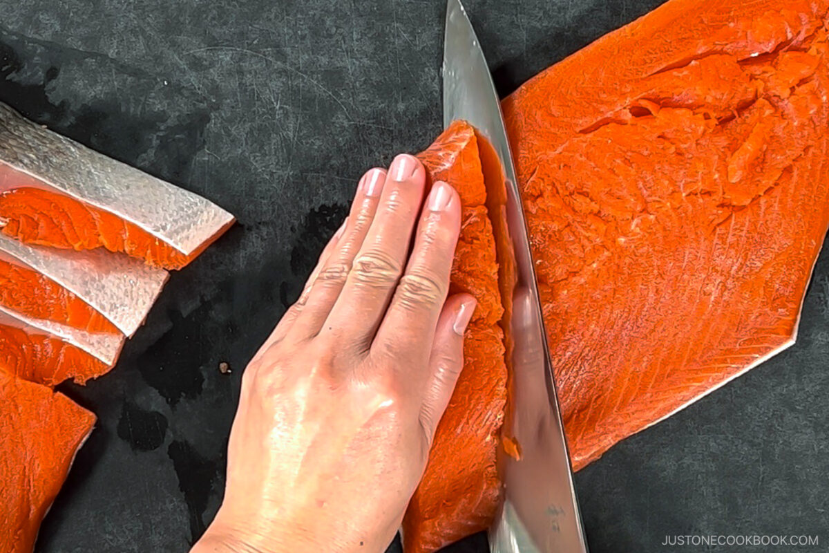 A hand holding a raw salmon fillet on a dark cutting board while slicing it with a large knife; several cut pieces are arranged to the left.