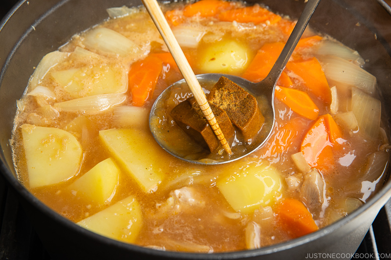 A pot of Japanese curry is simmering with chunks of potatoes, carrots, onions, and meat. A ladle with curry roux blocks—perfect for those learning how to make Japanese curry roux—and a pair of chopsticks are held over the bubbling broth.